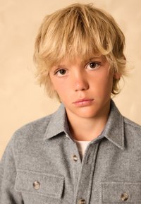 Chemise à chevrons grise à boutons, avec poches poitrine, ornée de boutons beiges, portée sur un T-shirt blanc, sur un fond neutre.