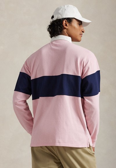 Férfi, rózsaszín hosszú ujjú ingben, amelyen széles tengerészkék csík fut a hátán, bézs nadrágban és fehér baseballsapkában, oldalra nézve.