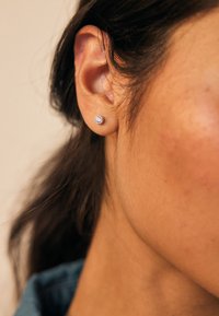 Pendiente de plata con un diseño circular y una gema transparente. El fondo muestra piel lisa y cabello oscuro, con el foco en el diseño del pendiente.