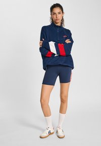 Felpa metà zip blu navy con maniche a righe rosse e bianche, abbinata a pantaloni ciclistici blu navy. Calze corte bianche e scarpe da ginnastica completano il look.