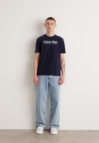 Marineblauwe katoenen T-shirt met witte "Calvin Klein" logo, gecombineerd met lichtblauwe denim jeans en witte sneakers, staand tegen een neutrale muur.