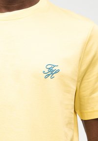 Κίτρινη κοντομάνικη μπλούζα με μικρό μπλε κεντημένο μονογράμμα "FH" στο αριστερό στήθος.