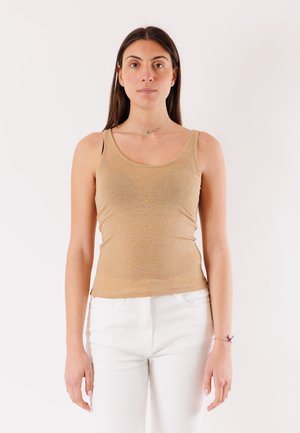 Donna con capelli lunghi e scuri che indossa un top beige senza maniche e pantaloni bianchi, in piedi davanti a uno sfondo bianco semplice.