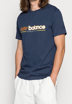 Hombre con camiseta azul marino de New Balance Athletics con logo blanco y naranja, combinada con pantalones blancos, manos en los bolsillos.