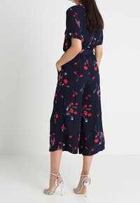 Tuta blu navy con stampa floreale rossa, maniche corte, vita aderente, design a gamba larga e tasche laterali. Indossata con sandali argentati con tacco alto.