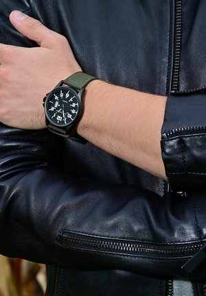 Black watch with large white numerals, green fabric strap, and black bezel, displayed on a wrist against a black leather jacket background.