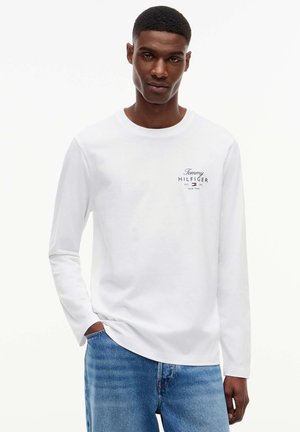 Man draagt een wit Tommy Hilfiger shirt met lange mouwen en een blauwe spijkerbroek, staand met één hand in de zak tegen een witte achtergrond.