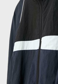 Nærbillede af en sort jakke med en vandret hvid stribe over brystet og en synlig sort lynlås i midten.