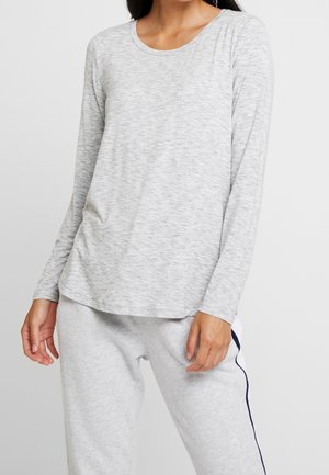 Kobieta ubrana w jasnoszary długi rękaw i dopasowane spodnie dresowe z granatowym paskiem po bokach, stojąca na tle jednolitego białego tła.