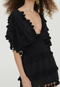 Černé letní šaty s hlubokým výstřihem do V, puffovými rukávy s černými třásněmi a nařaseným páskem s vrstvenými volány.