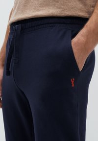 Donkerblauwe sweatpants met een elastische tailleband en trekkoord, voorzien van een klein rood embleem. Zijzakken zijn zichtbaar.