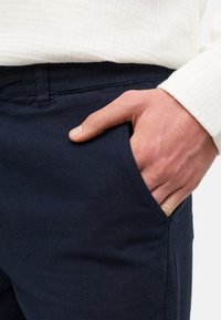Mornarsko plavi pamučni hlače s prednjim džepom; glatka tekstura; nosi se uz bijelu teksturiranu košulju. Ruka odmarajući u džepu, prikazuje detalje tkanine.