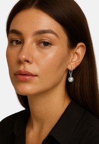 Pendientes de aros de plata con una gran perla blanca, llevados por una persona con cabello lacio y oscuro y maquillaje natural, sobre un fondo blanco.