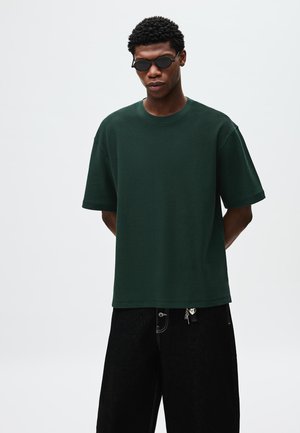 Tamnozelena majica kratkih rukava od teksturiranog materijala, s okruglim izrezom i opuštenim krojem, uparen s crnim širokim hlačama s detaljem cipela.