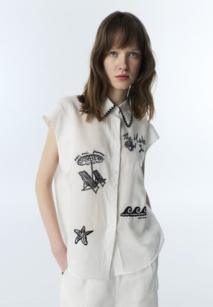 Jeune femme portant une chemise blanche sans manches avec une broderie noire sur le thème de la plage, incluant un parasol, des chaises, des vagues, une étoile de mer et du texte.