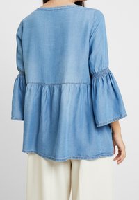 Light blue denim top featuring a flared sleeve design, gathered waist, and a soft texture. The hem is slightly curved, adding flow to the silhouette.
