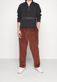 Sudadera negra con cremallera, bolsillo frontal y puños acanalados, combinada con pantalones de pana marrón de corte relajado y bolsillos laterales.