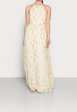 Woman wearing a sleeveless, floor-length white dress with yellow floral print and beige open-toe sandals, standing against a plain light background.