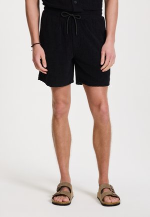 Zwarte gestructureerde shorts met een elastische tailleband en trekkoord. Draagt bij bruine sandalen met twee verstelbare banden.