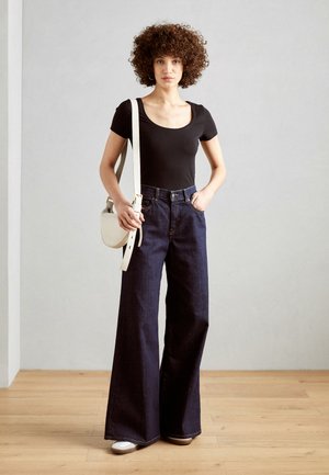 Top negro de escote redondo, jeans de mezclilla anchos de color azul oscuro, bolso cruzado blanco y zapatos blancos con acentos marrones, todo en un fondo neutro.