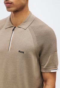 Homme portant un polo en maille beige à manches courtes avec épaules texturées, col zippé et logo noir "BOSS" sur la poitrine.