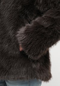 Σκούρο καφέ παλτό από συνθετική γούνα με μαλακό, υφασμάτινο φινίρισμα, διαθέτει εφαρμοστά μανίκια με ενσωματωμένη τσέπη. Casual τζιν από κάτω.