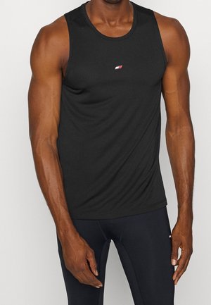 Personne musclée portant un débardeur noir ajusté sans manches et des leggings de sport noirs sur un fond uni.