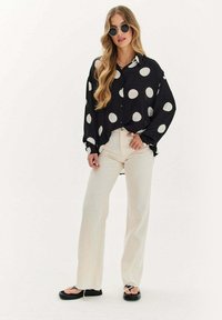 Czarna i biała bluzka w groszki o luźnym kroju, połączona z kremowymi spodniami w stylu dzwonów i czarnymi płaskimi sandałami. Strój ma swobodny wygląd.