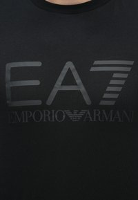 Fekete póló fényes "EA7" nagy betűkkel, alatta az "EMPORIO ARMANI" felirattal, a márka sas logójával a szavak között.