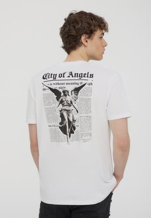 T-shirt bianca a maniche corte con una stampa grafica nera sul retro, che mostra un angelo circondato da testo di giornale.