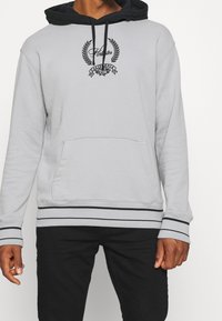 Sudadera con capucha gris con acentos negros, cordones ajustables, bolsillo frontal y logo bordado. Dobladillo y puños acanalados con rayas.