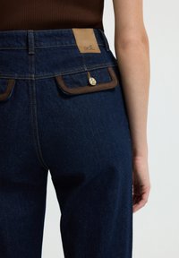 Pantalones de mezclilla azul oscuro con costuras claras, detalles de faux cuero marrón en los bolsillos y un parche con logo en la cinturilla. Incluye un botón dorado.