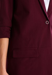 Bordeaux blazer met een getailleerde pasvorm, soepele textuur, enkele knoopsluiting en een discreet voorvak.