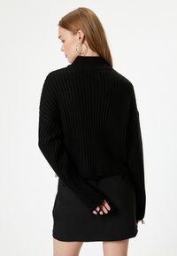 Schwarzer gerippter Strickpullover mit einem verkürzten Design, das einen hohen Kragen, einen ausgefransten Saum und ausgeschnittene Ärmel aufweist, kombiniert mit einem schwarzen Rock.