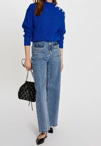 Niebieski sweter dzianinowy z metaliczną nitką, z guzikami na ramionach. Jasnoniebieskie dżinsy o wysokim stanie z luźnym krojem. Czarna torba typu bucket.