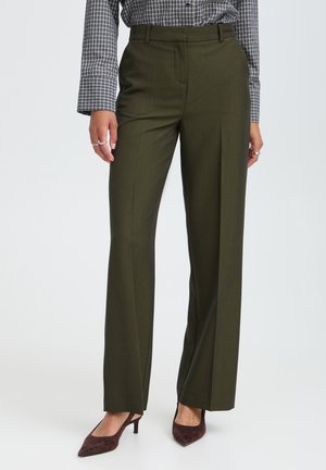 Personne portant un pantalon habillé vert olive sur mesure, une chemise grise à carreaux et des chaussures marron à talon bas, debout devant un fond uni.