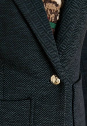 Tumši zaļš zīda jaciņa ar zelta pogu ar emblēmu un divām priekšējām kabatām, uzsverot teksturētu vilnas līdzīga materiāla. 