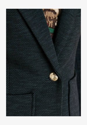 Tumši zaļš zīda jaciņa ar zelta pogu ar emblēmu un divām priekšējām kabatām, uzsverot teksturētu vilnas līdzīga materiāla.