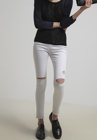 Vita rivna skinny jeans med fransar, kombinerat med en svart texturerad topp och svarta snörskor, hållande av en hand i en avslappnad pose.