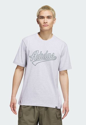 Giovane uomo con capelli biondi corti che indossa una maglietta Adidas grigio chiaro e pantaloncini cargo verde oliva, in piedi su uno sfondo semplice.