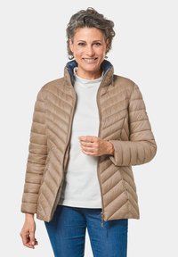 Giacca imbottita color marrone chiaro con colletto alto e chiusura con zip, abbinata a un top bianco e jeans blu. Tessuto liscio, design aderente.