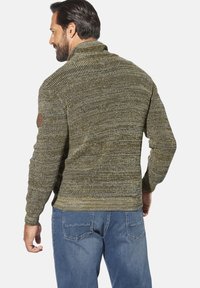 Mann in olivgrünem Strickpullover und blauen Jeans, mit dem Rücken zur Kamera und entspannten Händen an den Seiten.