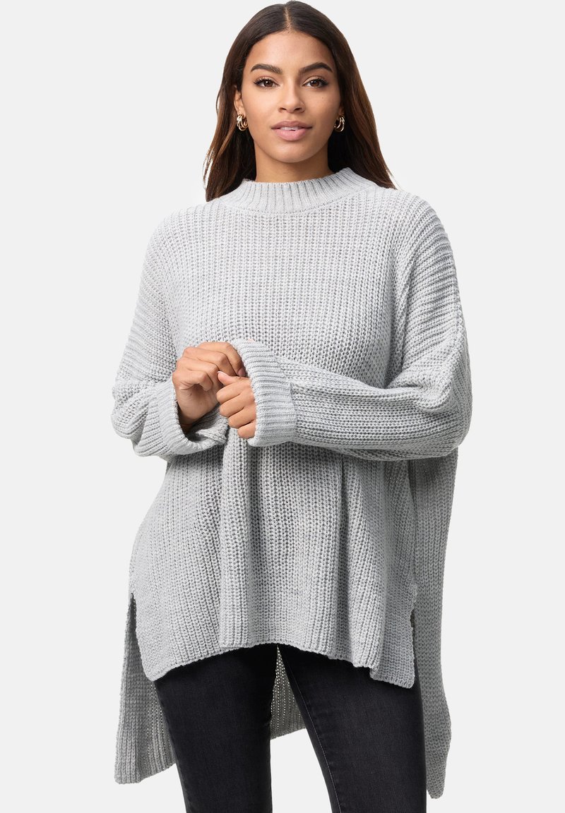 Hellgrauer, übergroßer Strickpullover mit hohem Kragen, geripptem Textur, seitlichen Schlitzen und überschnittenen Schultern, kombiniert mit dunklen Jeans.