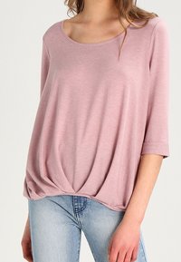 Helles Pink, lässiges Oberteil mit Rundhalsausschnitt und drei Viertelärmeln. Das Material wirkt weich und hat eine Raffung am Saum.