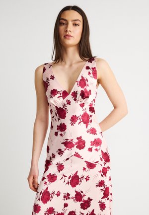 Frau trägt ärmelloses, hellrosa Kleid mit tiefem V-Ausschnitt und rotem Blumenmuster, steht vor einfachem weißem Hintergrund.