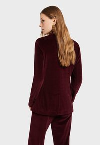 Burgundský velúrový kabát s dlhými rukávmi, rovný strih a detailom švu na chrbte. Hladká textúra a kombinovaný s ladnými nohavicami.