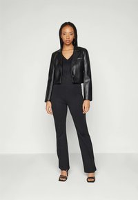 Veste en cuir noire coupée sur un haut noir et un pantalon noir ajusté. Modèle se tenant debout avec les mains sur les hanches, portant des talons aiguilles noirs.