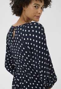 Robe bleu marine à pois avec une ouverture en forme de goutte dans le dos, un col froncé et des manches longues. Le tissu semble léger avec une texture lisse.