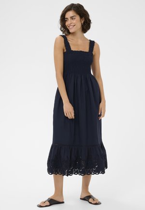 Femme portant une robe midi bleu marine sans manches avec des bretelles volantées et un ourlet ajouré, associée à des sandales plates noires, debout et souriante.