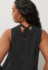 Top senza maniche nero con chiusura legata nella parte posteriore, leggermente drappeggiato e con una texture liscia. Presenta un'apertura a chiave sul retro e arricciature sulle spalle.